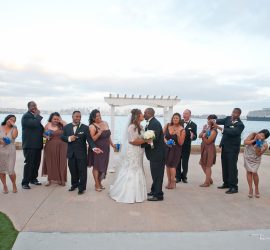 A bride and groom kiss outdoors as their wedding party reacts with playful laughter and surprise near a waterfront.