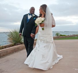 A bride and groom walk hand in hand outdoors by the water, smiling at each other in wedding attire.