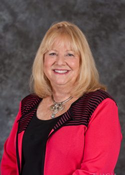 Smiling woman with long blonde hair wearing a bright pink jacket and statement necklace, posing in front of a gray backdrop.