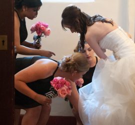 A bride in a white gown adjusts her shoe, assisted by three bridesmaids holding pink bouquets in a small room.