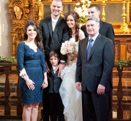 Six adults and one child pose together in formal attire in front of an ornate altar at a wedding ceremony.