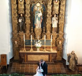 A bride and groom stand together in front of an ornate church altar decorated with statues and candles.