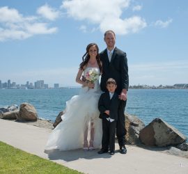 Bride, groom, and young boy pose together by a waterfront with city skyline in the background on a sunny day.