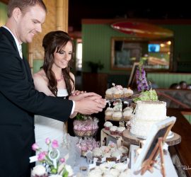 A bride and groom cut a wedding cake together at a decorated table with cupcakes and flowers.