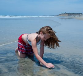 Young girl in a swimsuit kneels on wet sand at the beach, touching the water under a blue sky.