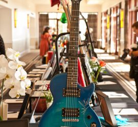 A signed blue Epiphone electric guitar on display at an indoor event, surrounded by flowers and framed photos.