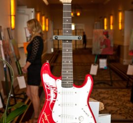 Red and white electric guitar with autographs on display at an indoor event; blurred people and artwork in background.