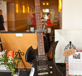 Black electric guitar with white signatures displayed upright on a table next to a rose in a vase and a framed sign labeled "2013.
