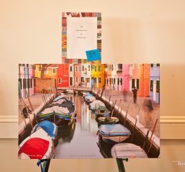 A photograph of boats moored along a canal with colorful buildings, displayed on an easel with a label above it.