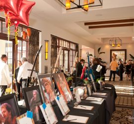 A silent auction table with framed photos and bids, guests mingle in a well-lit event space with red star balloons.