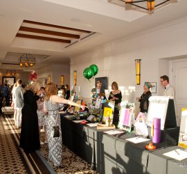 People view and discuss items displayed on tables at an indoor event with bright lighting and decorative balloons.