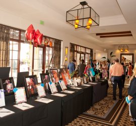 People browse a silent auction display with bid sheets and photos in a well-lit hallway during an event.
