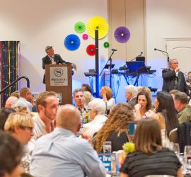 A man speaks at a podium to an audience seated at tables in a decorated banquet hall with stage equipment visible.