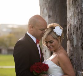 A couple in wedding attire stands by a tree; the groom rests his forehead on the bride’s head as she holds red roses.