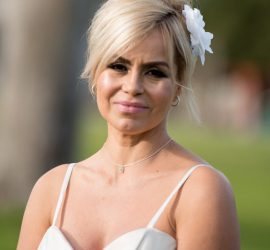 A woman in a white dress stands outdoors, smiling, with a flower in her hair and blurred greenery in the background.