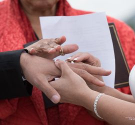 A bride places a ring on the groom’s finger during a wedding ceremony, with an officiant holding papers in the background.