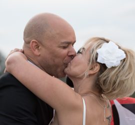 A bride and groom share a kiss outdoors, with the bride wearing a white flower in her hair.