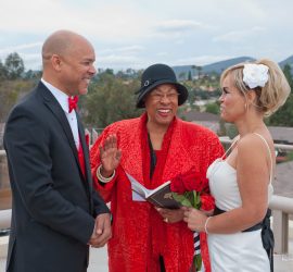 An officiant in red stands between a couple exchanging vows outdoors, with hills and rooftops in the background.