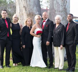 A wedding group portrait with seven people standing outdoors in formal attire, with two large trees in the background.