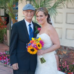 A bride in a white dress holds a bouquet, standing next to an older man in a suit and flat cap, outdoors.