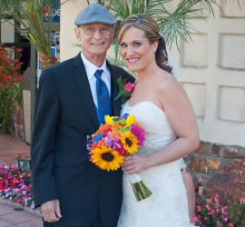 A bride in a white dress holds a bouquet, standing next to an older man in a suit and flat cap, outdoors.