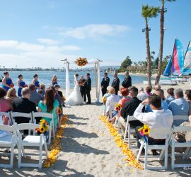 Outdoor beach wedding ceremony with guests seated, couple at the altar, and sailboats on the water in the background.