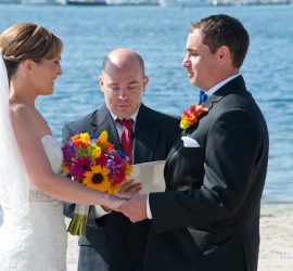 A bride and groom hold hands and face each other during an outdoor wedding ceremony by the water with an officiant.