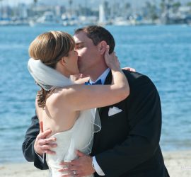 A bride and groom kiss by the water, dressed in wedding attire, with boats and palm trees in the background.