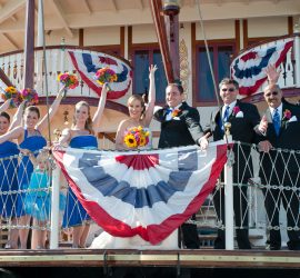 A wedding party in formal attire poses and waves on the deck of a boat decorated with red, white, and blue bunting.
