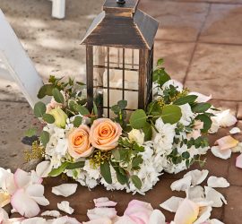 A black lantern with a candle inside is surrounded by peach roses, white flowers, and scattered pink rose petals.