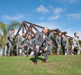 A group of men in gray suits pose on a grassy hill in front of a wooden structure under a blue sky.