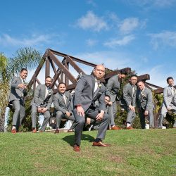A group of men in gray suits pose on a grassy hill in front of a wooden structure under a blue sky.