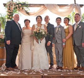 Seven people posing for a formal group photo at an outdoor wedding ceremony with floral decorations.