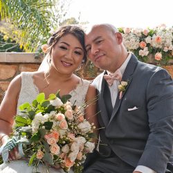 A bride and groom sit closely together, smiling, holding a bouquet of white and peach roses, with floral decor behind them.