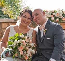 A bride and groom sit closely together, smiling, holding a bouquet of white and peach roses, with floral decor behind them.