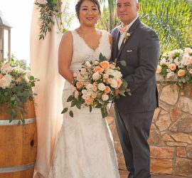 A bride and groom stand together, smiling, in front of a floral wedding arch and stone wall decorated with peach roses.
