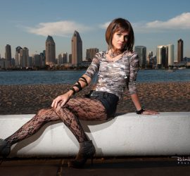 A woman in patterned tights and heels sits on a low wall by the water with a city skyline in the background.
