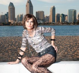 A woman in patterned tights and heels poses in front of a waterfront city skyline under a clear sky.