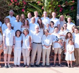 A large multigenerational family poses outdoors on steps, all dressed in white and beige clothing with greenery behind them.