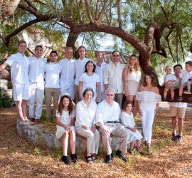 A large family dressed in white poses outdoors under a leafy tree, with adults standing and children sitting or perched on laps.