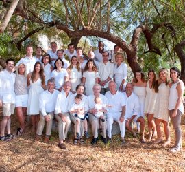 A large family group poses outdoors under a tree, all wearing white or light-colored clothing.
