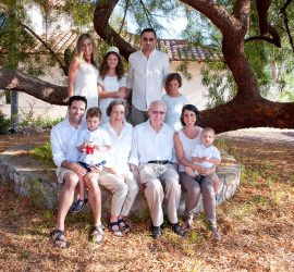 Three generations of a family pose outdoors under a large tree, dressed in light-colored, casual clothing.