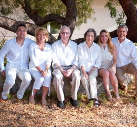 Six people sit on a stone bench under a tree, all wearing white shirts and smiling at the camera.