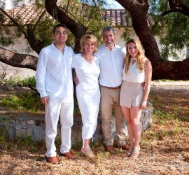 Four people pose together outdoors in front of a large tree, all smiling and dressed in light-colored clothing.