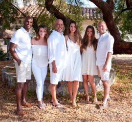 Six adults stand outdoors under a large tree, all dressed in white, smiling at the camera.