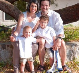 A family of four poses outdoors in front of a tree, smiling at the camera on a sunny day.