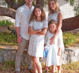 A family of four poses outdoors in front of a tree and stone wall, all dressed in light-colored clothing.