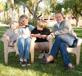 A family of four sits together on lawn chairs outside, smiling and interacting in a grassy yard with trees.