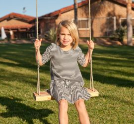 Young child in a striped dress sits on a wooden swing outside on a sunny day with houses and palm trees in the background.