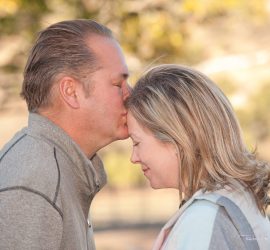 A man gently kisses a woman's forehead outdoors as both stand with their eyes closed.